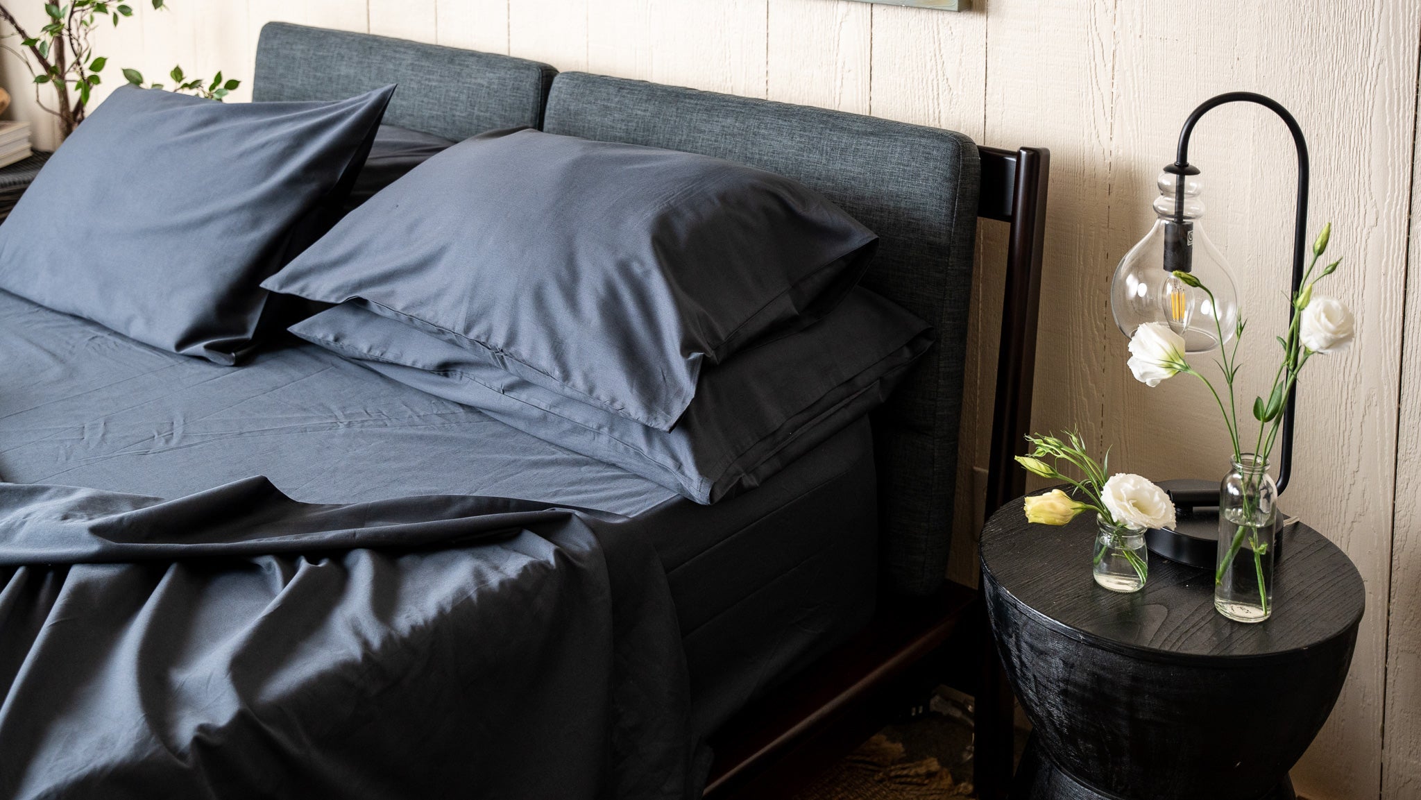 Bed with dark gray bedding and pillows, nightstand with flowers and lamp.
