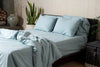 Bed with light blue bedding and pillows in a room with plants and a wooden shelf.
