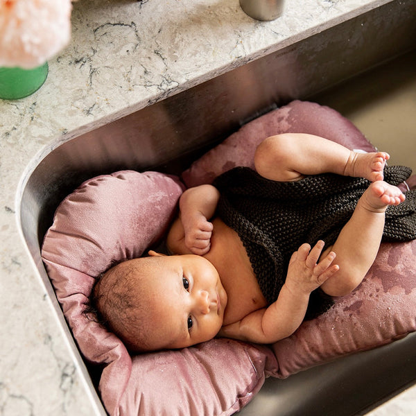 Plum+Sparrow Infant Sink Bather in Orchid Haze shown in a standard kitchen sink, supporting a newborn baby during bathtime