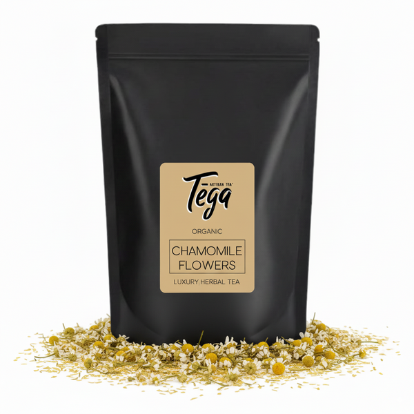 Black packaging of Tega Organic Chamomile Flowers tea with scattered chamomile flowers on a white background
