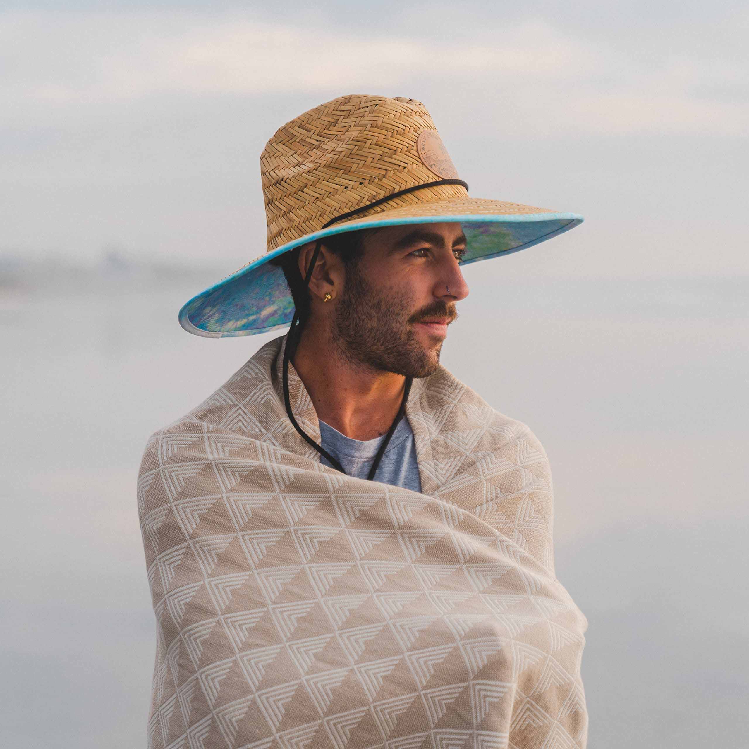 Man wearing a straw hat and wrapped in a patterned blanket against a blurred natural background