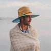 Man wearing a straw hat and wrapped in a patterned blanket against a blurred natural background