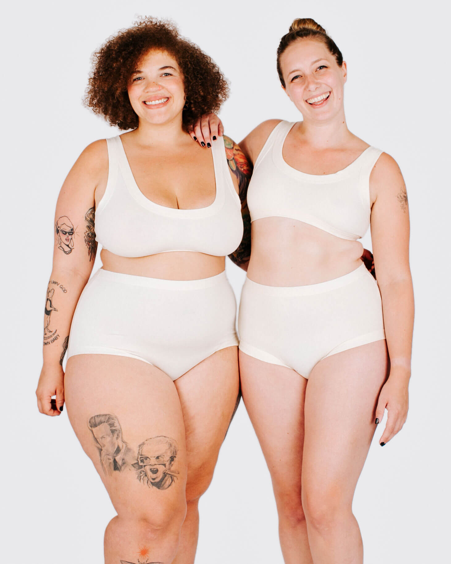 Two women wearing white tank tops and high-waisted underwear standing side by side on a plain background.