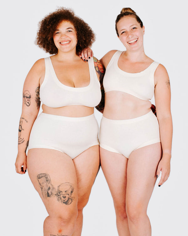 Two women wearing white tank tops and high-waisted underwear standing side by side on a plain background.