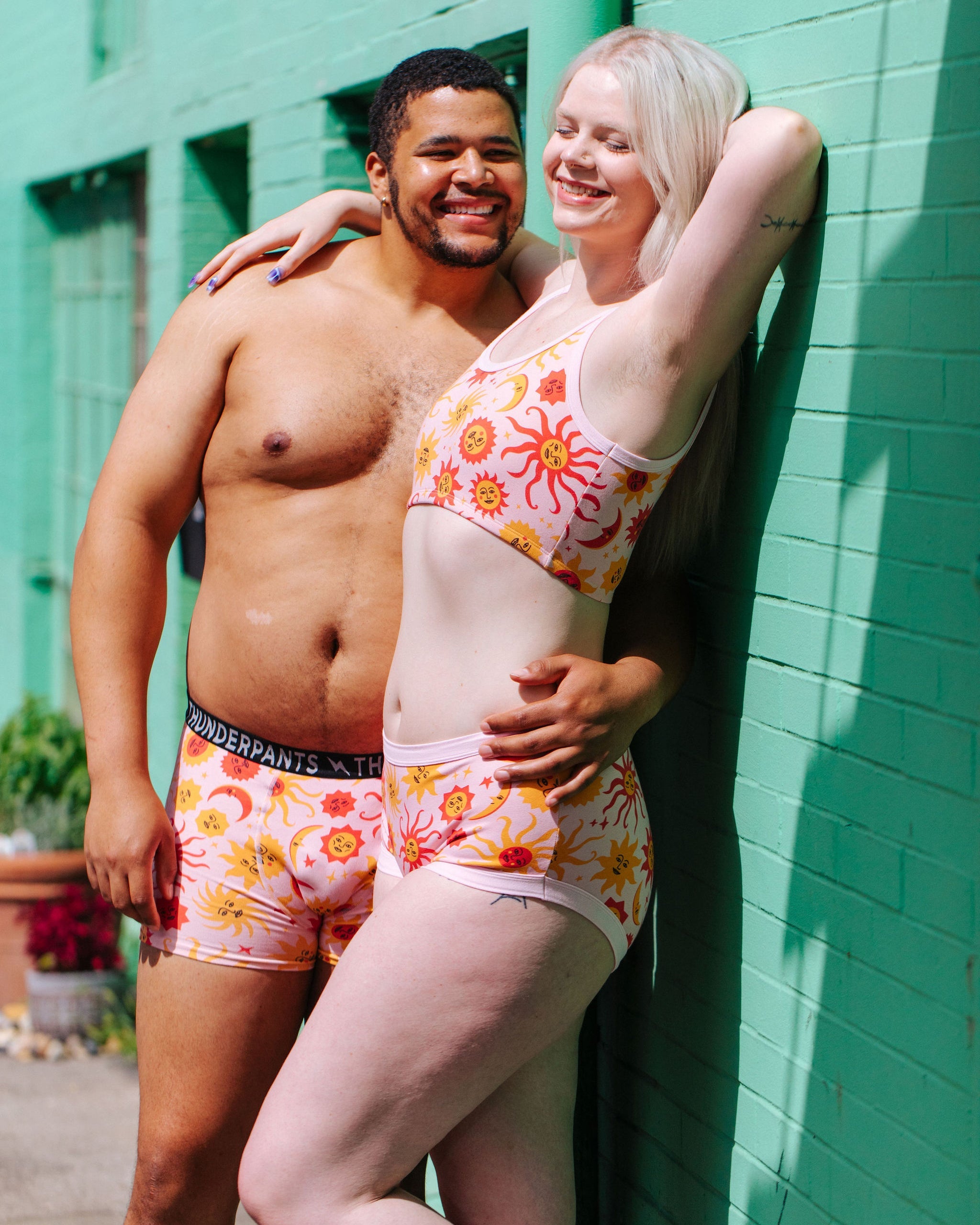 Two people wearing matching colorful underwear in front of a green wall.
