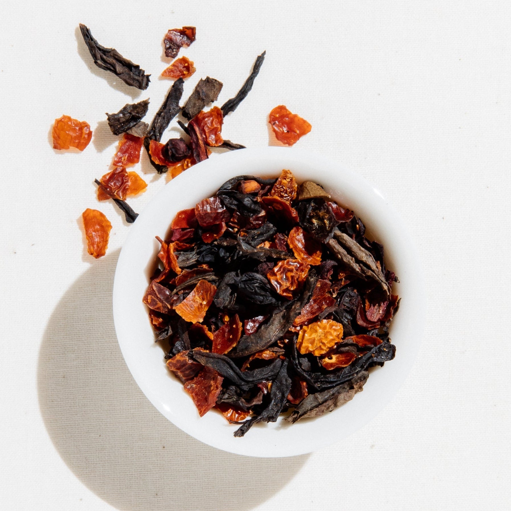 Loose leaf Plum Oolong tea by Art of Tea in a small white bowl, showing dried amber oolong leaves, schizandra berries, and bright orange rosehips on a white background