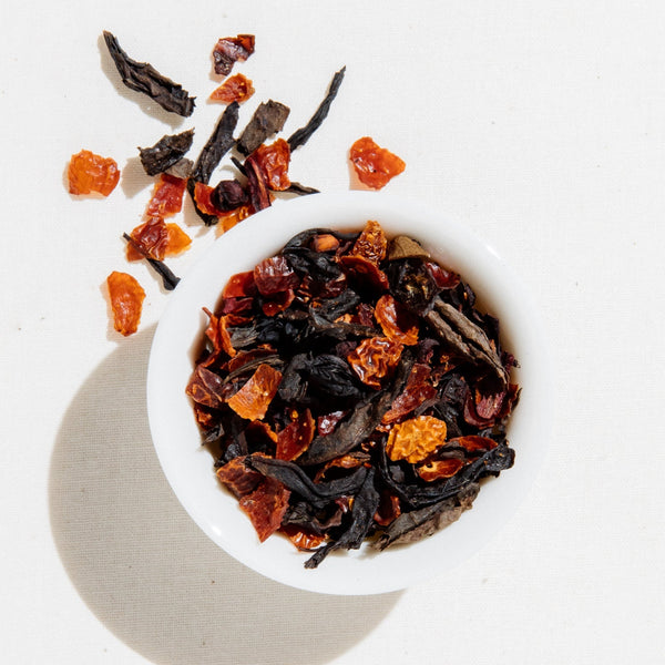 Loose leaf Plum Oolong tea by Art of Tea in a small white bowl, showing dried amber oolong leaves, schizandra berries, and bright orange rosehips on a white background