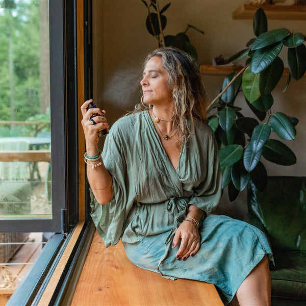 Woman in a green dress sitting by a window with plants around her using paavani rose water mist