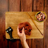 Hand holding a piece of meat on a wooden cutting board with a small bowl of additional meat and measuring spoons.