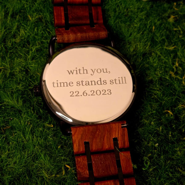A wooden watch with a black dial and brown strap, displayed on a natural wood surface.