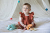 Rust muslin baby romper styled with orange teething rattle, stroller toy, and first baby doll