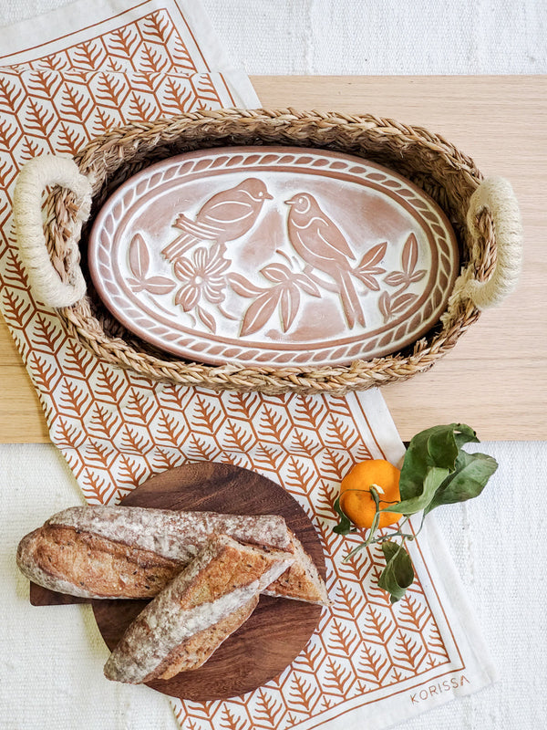 Decorative plate with bird design in a woven basket on a table with bread and an orange.