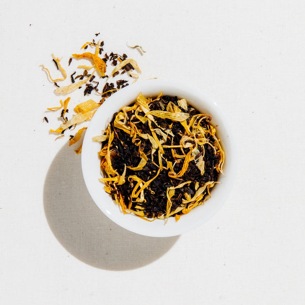 Dried loose-leaf tea in a small white bowl with a few loose strands scattered beside it, photographed from above on a white background.
