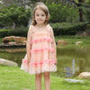 A young girl stands outdoors in a grassy park area with a pond and trees in the background, wearing a light peach pink tiered tulle dress with ruffled trim and a matching headband.