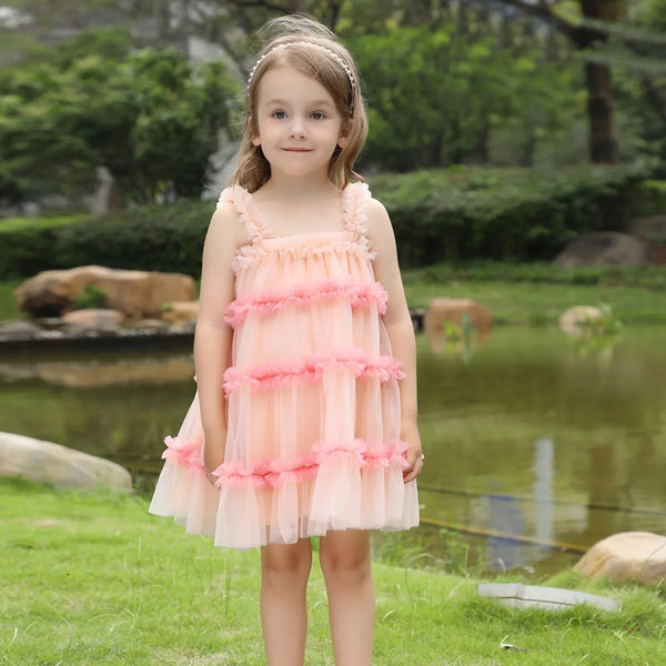A young girl stands outdoors in a grassy park area with a pond and trees in the background, wearing a light peach pink tiered tulle dress with ruffled trim and a matching headband.
