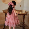 Young girl in a pink dress sitting at a wooden table with a lamp and candle holder in the background.