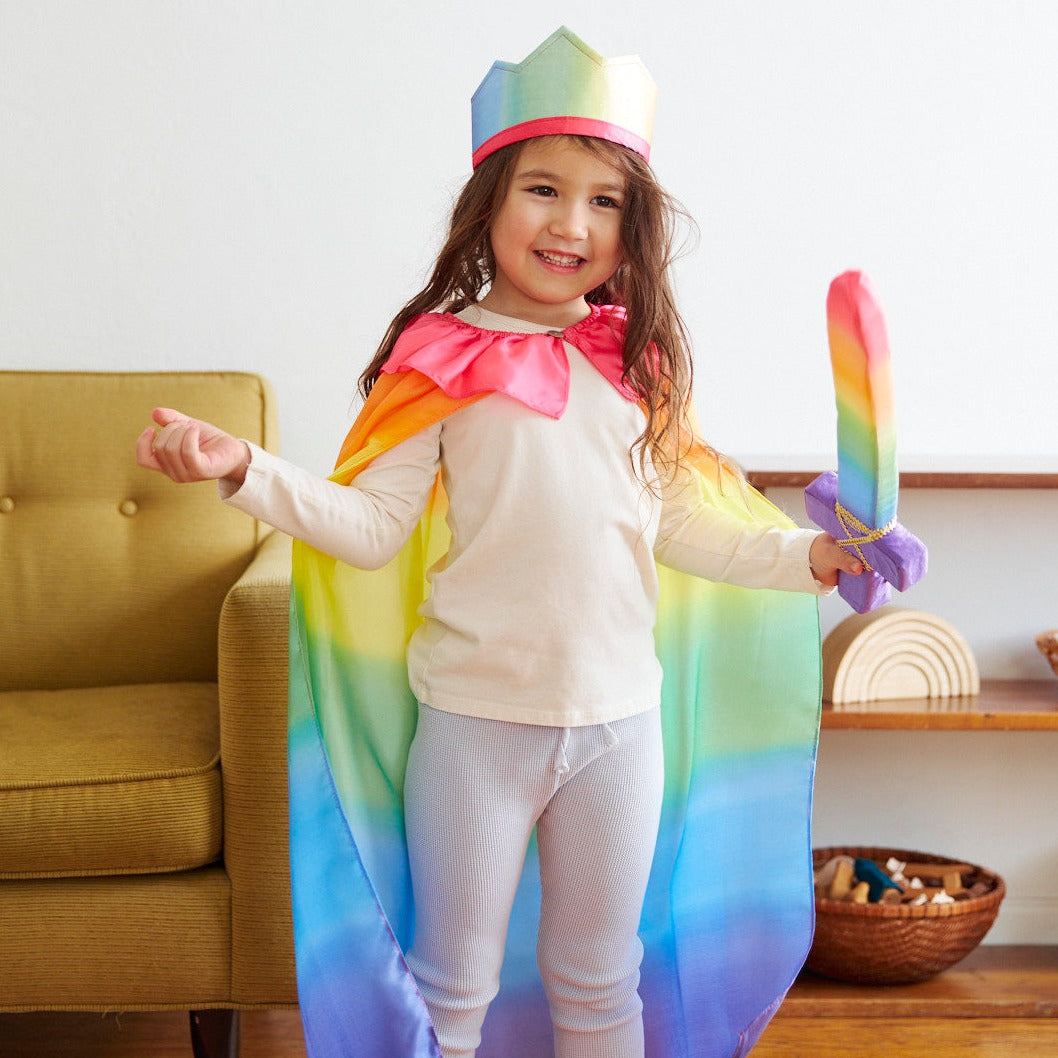Child in a rainbow-themed costume with a cape and crown, standing in a room with a couch and shelves.
