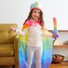 Child in a rainbow-themed costume with a cape and crown, standing in a room with a couch and shelves.