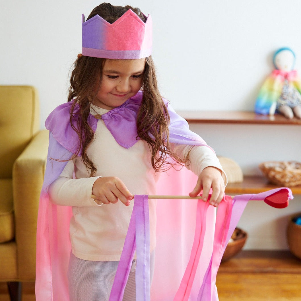 Child wearing a colorful cape and crown in a room with a couch and shelves.