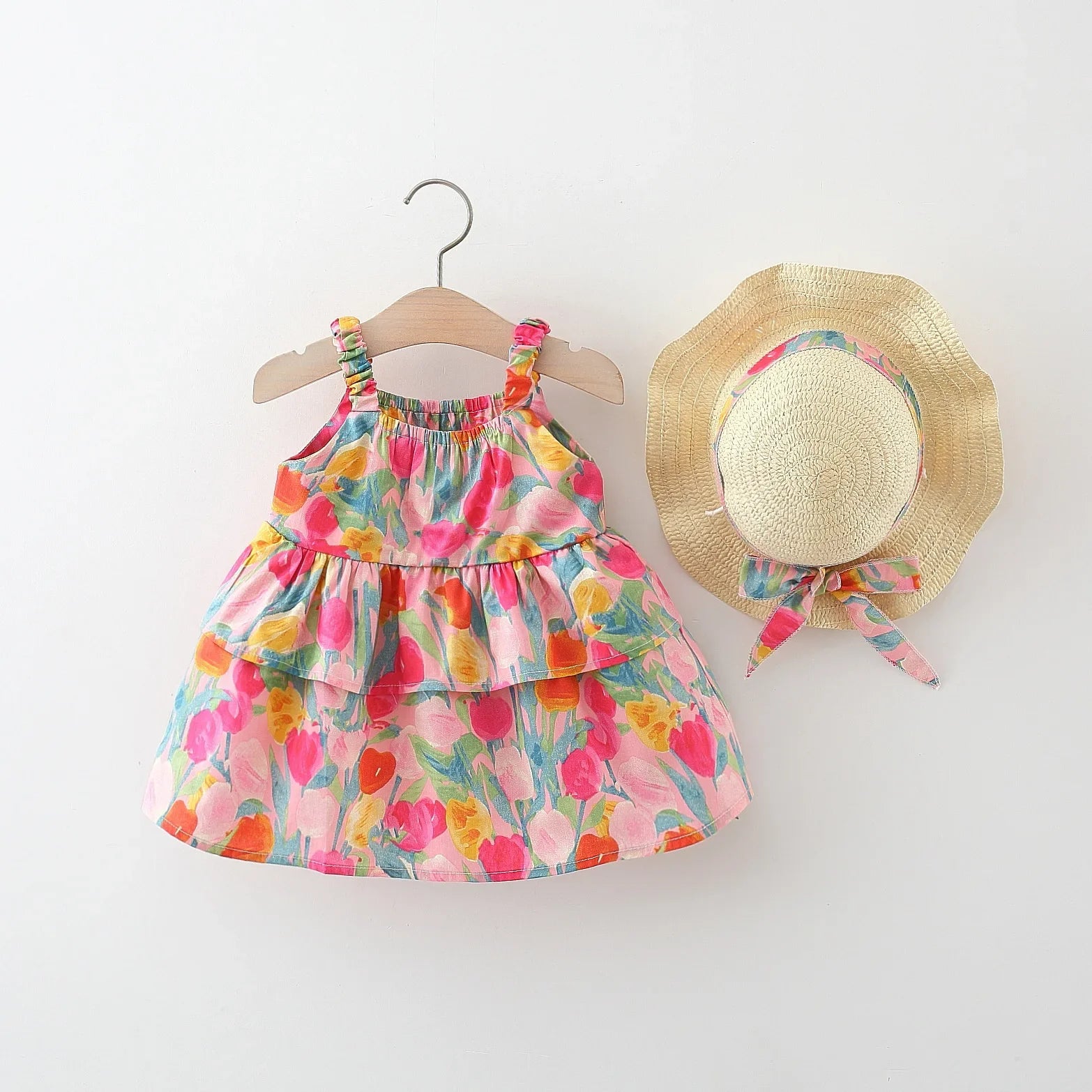 A toddler girl's sleeveless tiered dress with a colorful floral tulip print, displayed on a wooden hanger next to a matching straw sun hat with a ribbon bow, against a clean white background.