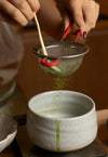 Person using a metal sieve to pour green powder into a ceramic bowl.