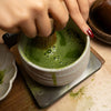 Person using a whisk to prepare matcha green tea in a ceramic bowl.