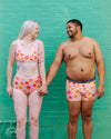 Two people wearing matching floral underwear in front of a teal brick wall.