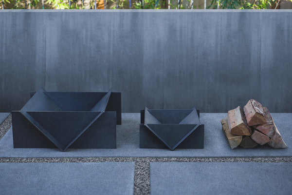 Two dark metal fire pits on a stone patio with stacked firewood against a gray wall.