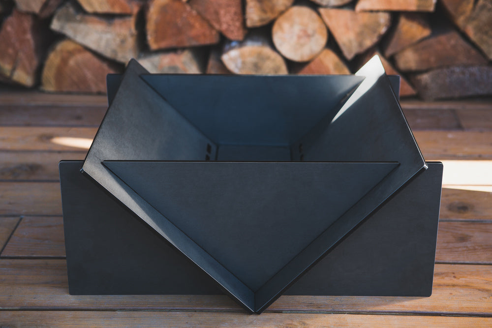 Black metal fire pit on a wooden surface with stacked firewood in the background