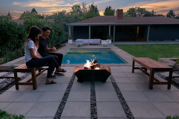 Couple sitting by a fire pit in a backyard with a pool and house in the background.