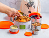 Person using a stainless steel pet food container with various food items and orange lids on a light background.