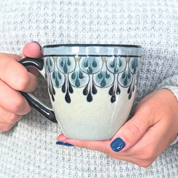 Handpainted Fair Trade stoneware coffee mug with black and blue Mayan geometric patterns, held against a textured surface