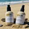 Apotheke DuJardin Sun Shield and Sun Glow reef safe tallow barrier oil bottles nestled together in beach sand with ocean waves in the background