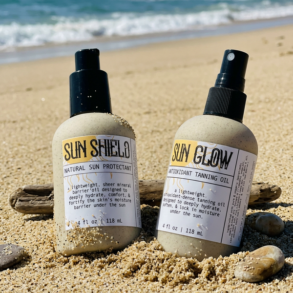 Apotheke DuJardin Sun Shield and Sun Glow reef safe tallow barrier oil bottles nestled together in beach sand with ocean waves in the background