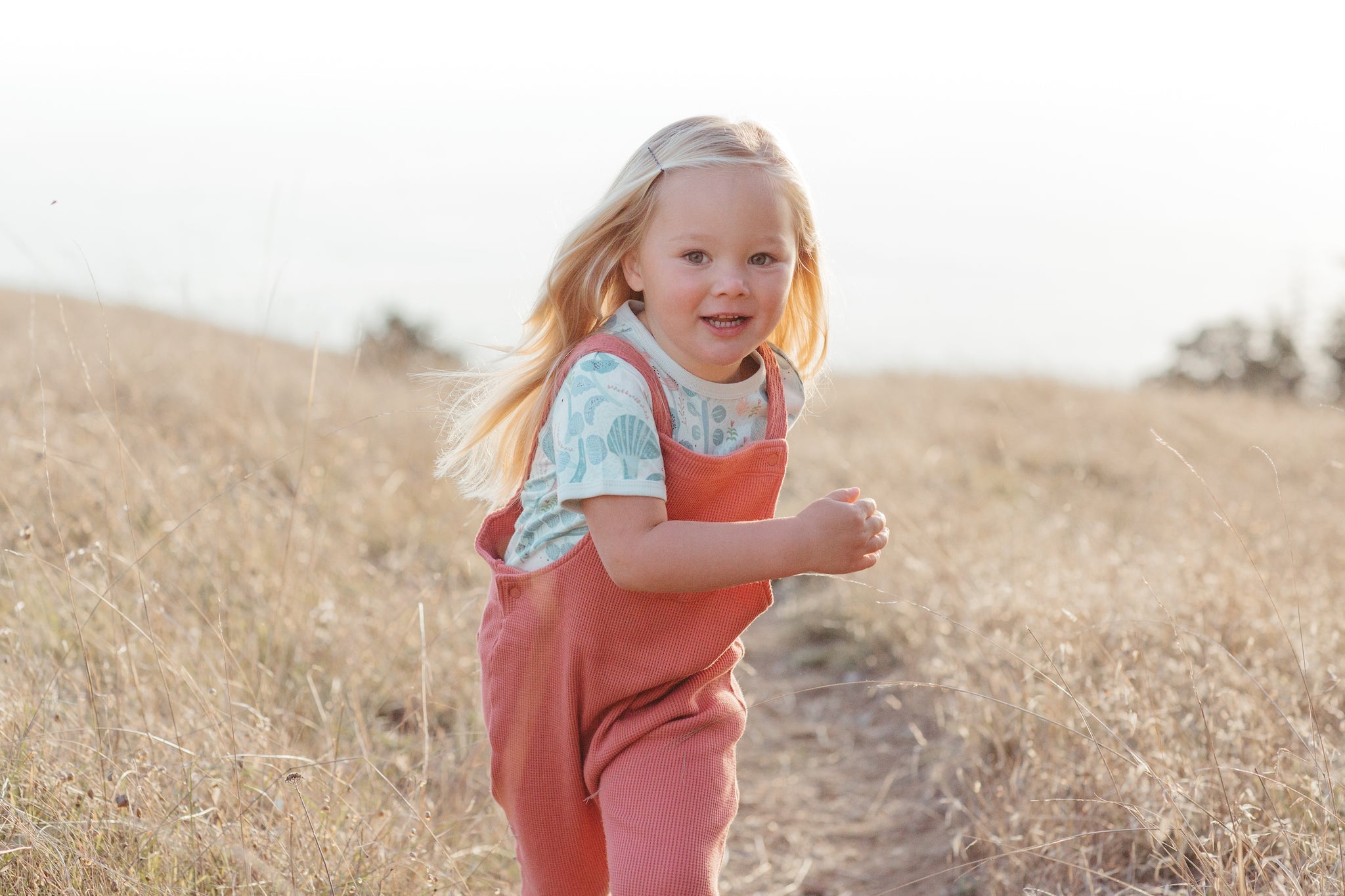 Child in pink overalls standing on a dirt path in a field
