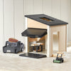 Children's playhouse with black roof and wooden floor, surrounded by toy cars and a wheelbarrow on a light wood floor.
