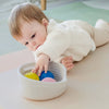Baby playing with colorful balls in a woven basket on a light-colored floor.