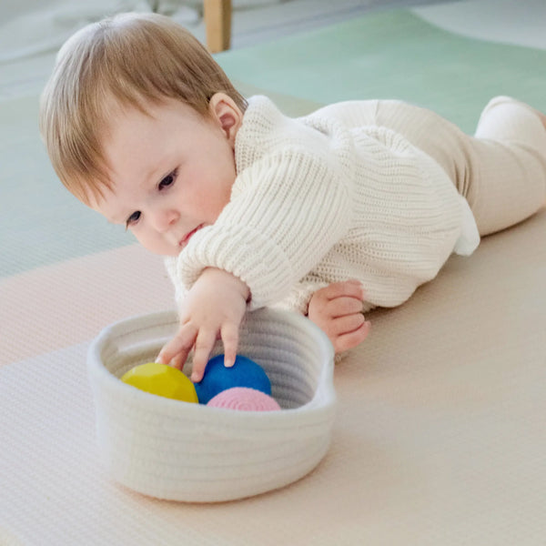 Baby playing with colorful balls in a woven basket on a light-colored floor.