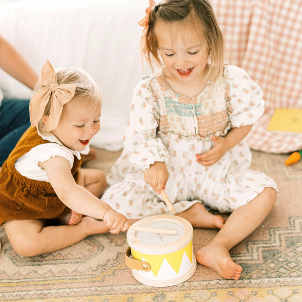 Two young children playing with a toy drum on a patterned rug.