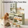A young girl with long light brown hair sits on the floor next to a sage green wooden play kitchen set. The kitchen features a stove, oven, sink with a gold faucet, shelves, and storage bins.