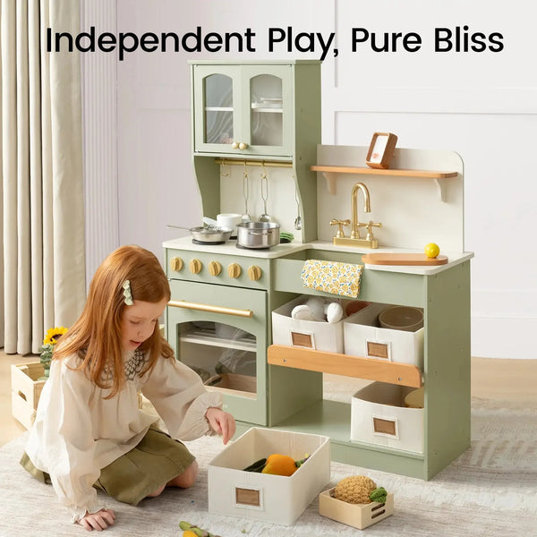 A young girl with long light brown hair sits on the floor next to a sage green wooden play kitchen set. The kitchen features a stove, oven, sink with a gold faucet, shelves, and storage bins.