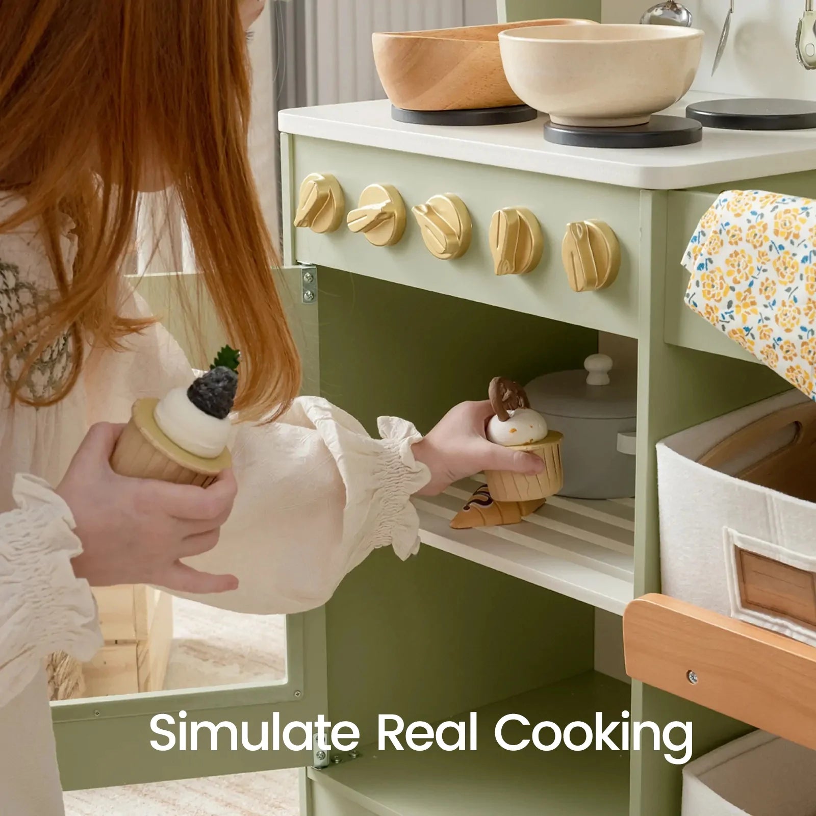 A child with long red hair plays with a wooden toy kitchen, placing a small toy dessert into the cabinet. The toy kitchen is sage green with cream-colored knobs and a white countertop, and the text 'Simulate Real Cooking' is overlaid at the bottom.