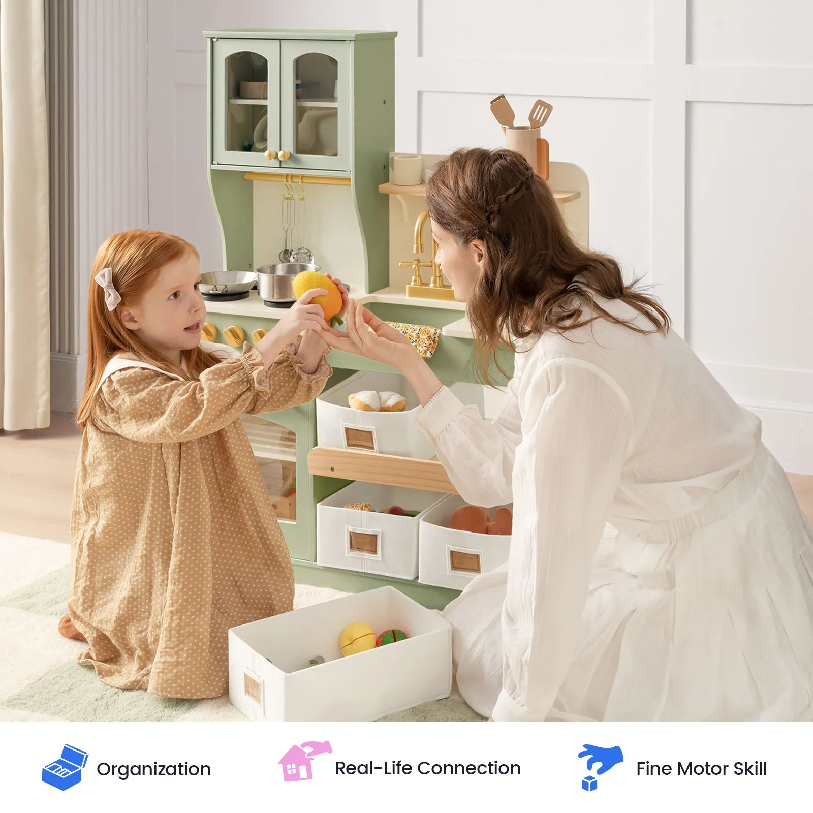A woman and a young girl play with a wooden toy kitchen set in a bright, modern room. The toy kitchen is light green with white storage bins and includes a sink, faucet, and small kitchen accessories.