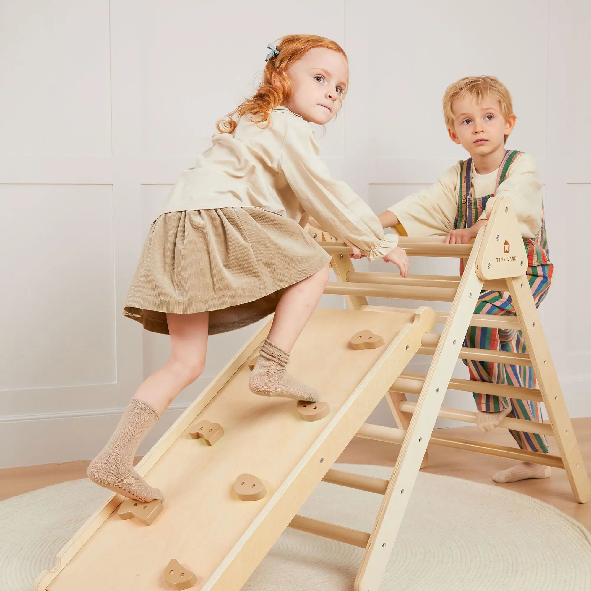 Two children playing on a wooden climbing toy against a white wall.