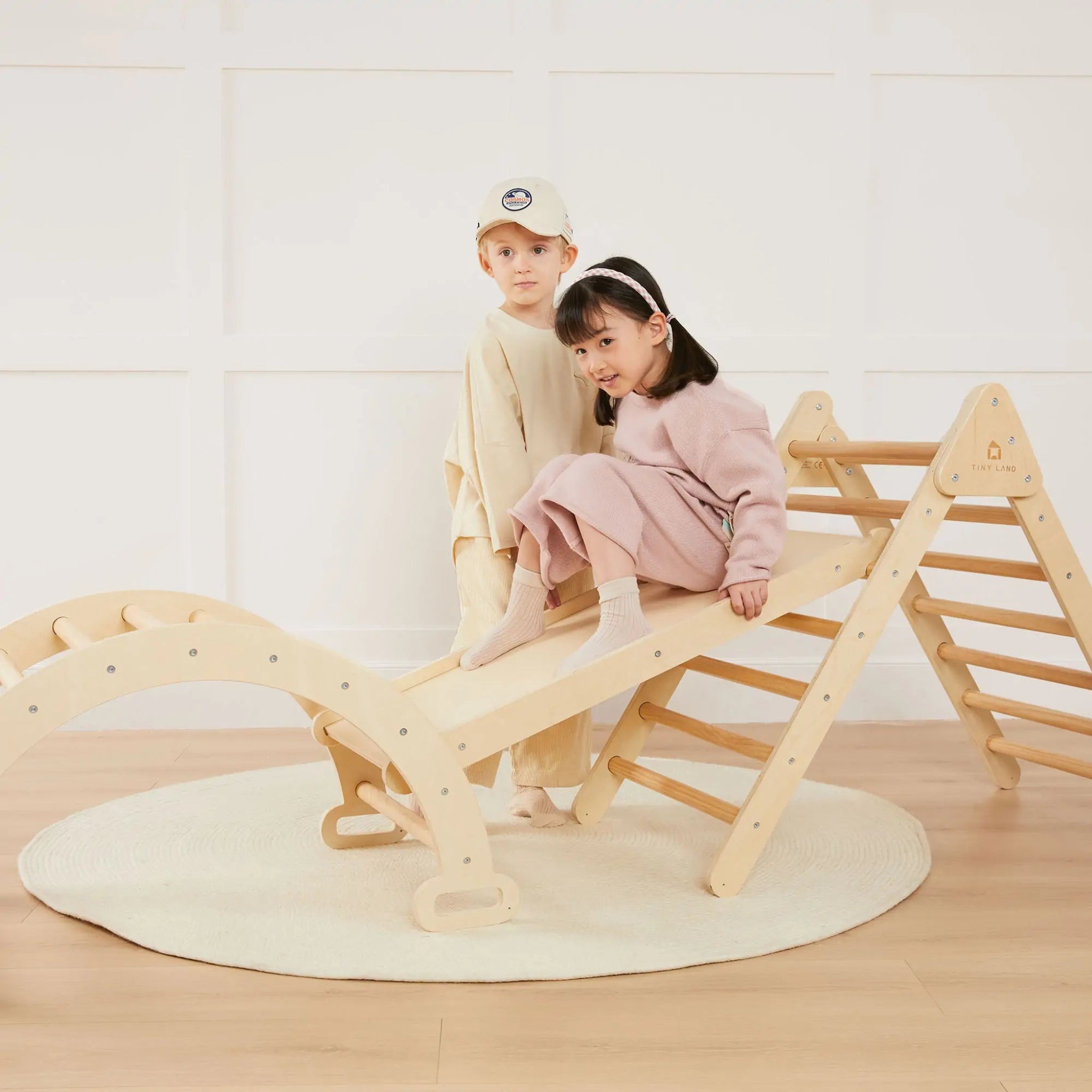 Two children playing on a wooden seesaw and ladder set indoors.