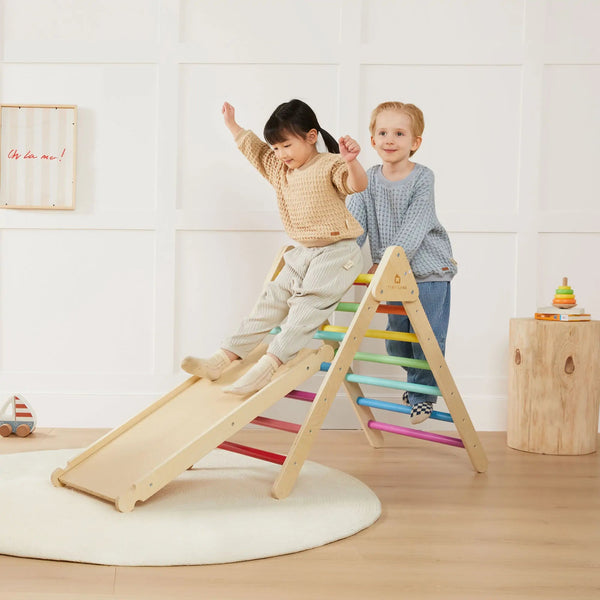 Two children playing on a colorful wooden climbing toy in a room with a white wall and wooden floor.