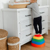 Child standing on a colorful step stool next to a white dresser with wicker baskets.
