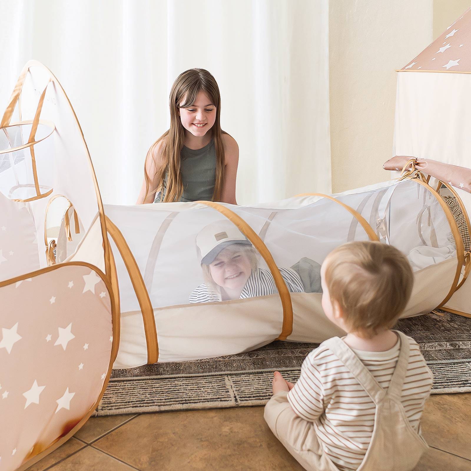 Two children playing with a tunnel toy in a room with white curtains.