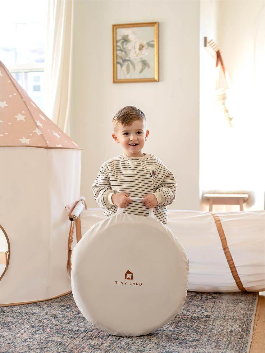 Child sitting on a beige ball with 'Tiny Land' logo in a room with play tent and decor.