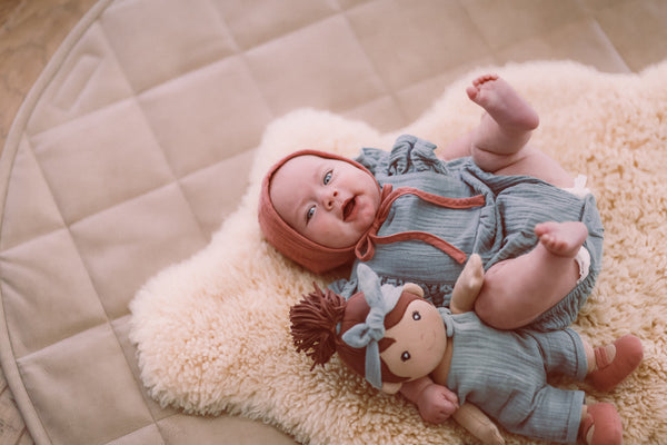 Baby wearing teal muslin flutter sleeve romper with terra cotta muslin bonnet and Paloma park friend soft toy